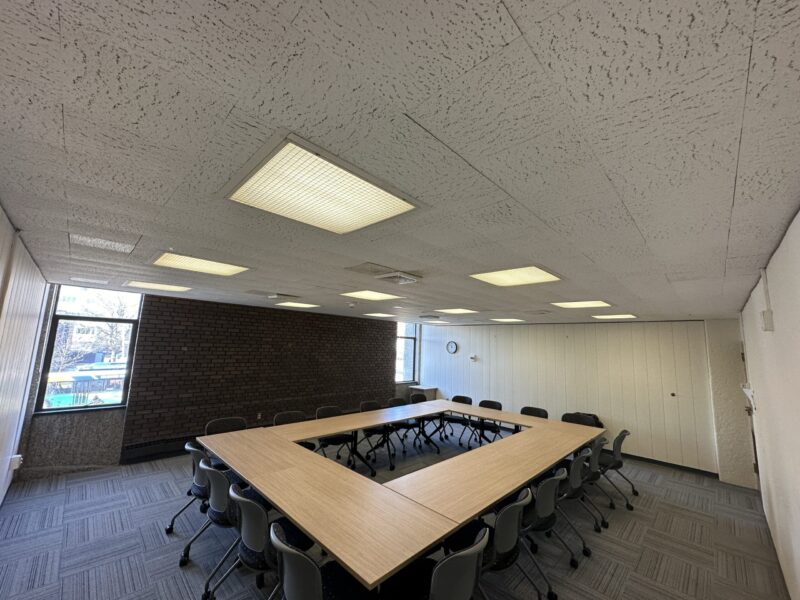 A conference room with U-shaped tables made of light wood, surrounded by gray chairs on wheels. The room has gray patterned carpet, white textured ceiling with recessed fluorescent lights, and beige paneled walls. Two large windows on the left and far wall provide natural light and show an outdoor view with trees and buildings. A small wall clock is mounted on the far wall.