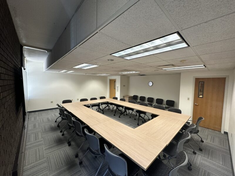 A conference room with U-shaped tables made of light wood, surrounded by gray chairs on wheels. The room has gray patterned carpet, white walls, and a white drop ceiling with fluorescent lighting panels. A wooden door with a small window is on the right wall, and another door is at the back. A small wall clock is mounted on the far wall above a row of extra chairs. One wall is dark brick, and a flat-screen monitor is partially visible on the left.
