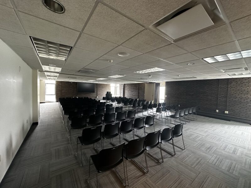 A large meeting or event room set up in auditorium style with multiple rows of black chairs facing a podium and a large wall-mounted screen at the front. The room has gray patterned carpet, a white drop ceiling with recessed fluorescent lights, and dark brick walls. Natural light enters through tall windows at the far end of the room.