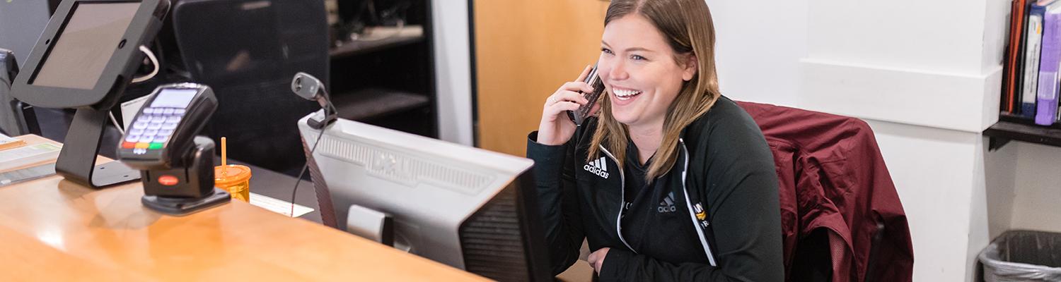 A front desk student employee smiling and talking on the phone at the Klotsche Center
