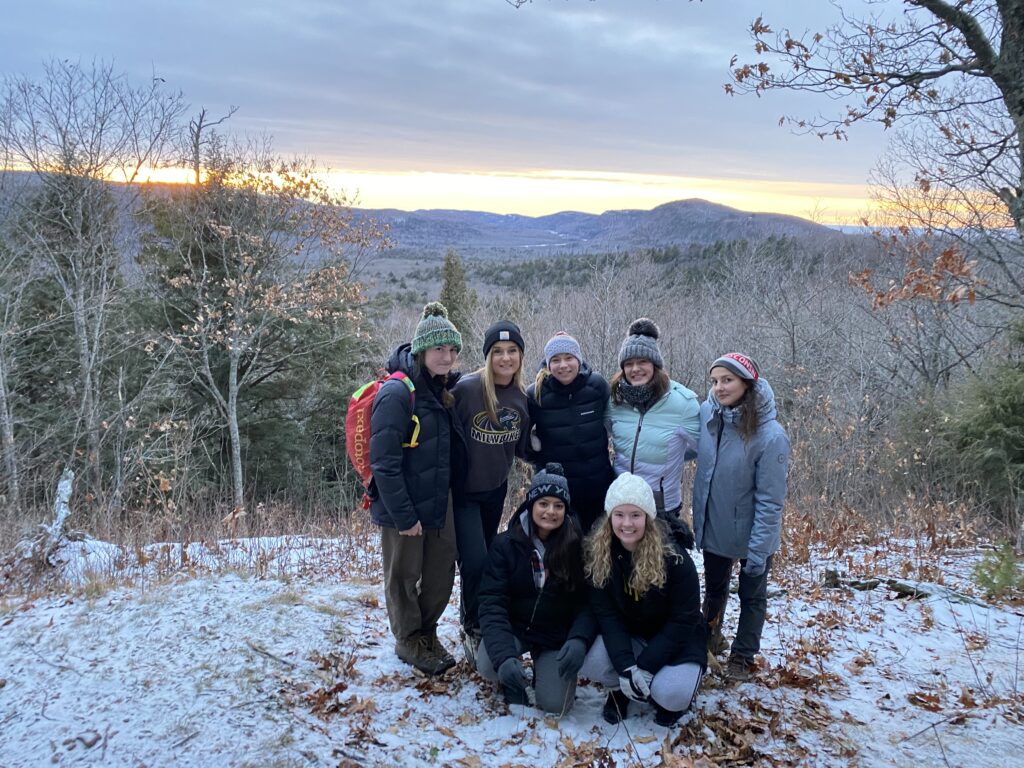two rows of women huddling at a snowy overlook a wooded hike in winter coats and hats