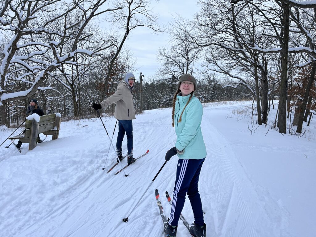 two people cross country skiing on a snowy, woodsy trail looking back at the camera smiling
