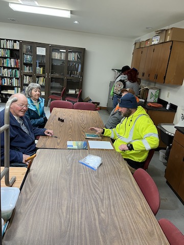 Group meeting in a library conference room with students and instructors seated around a wooden table, bookshelves visible in background.