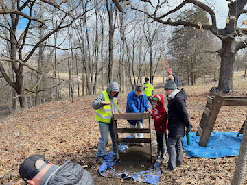 Students in safety vests gather around equipment for outdoor field research in a winter forest setting.