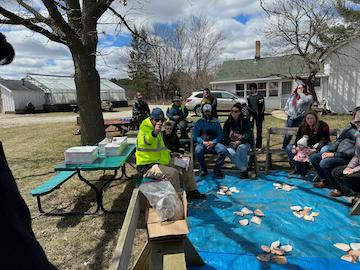 Students gathered around a blue tarp with research materials spread out, conducting outdoor field work near campus buildings under partly cloudy skies.