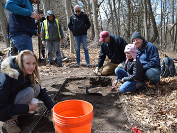 Students conducting archaeological or soil research in a forest, with some kneeling to examine the ground while others observe, orange bucket visible.
