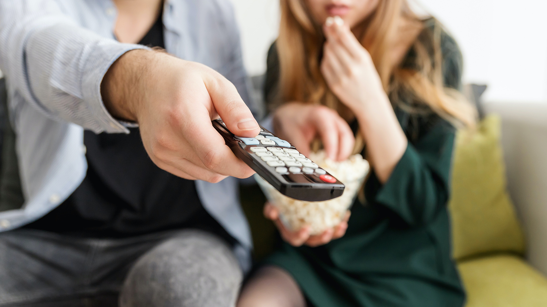 A photo of a man and a woman enjoying popcorn from the neck down while turning the TV channel on the remote