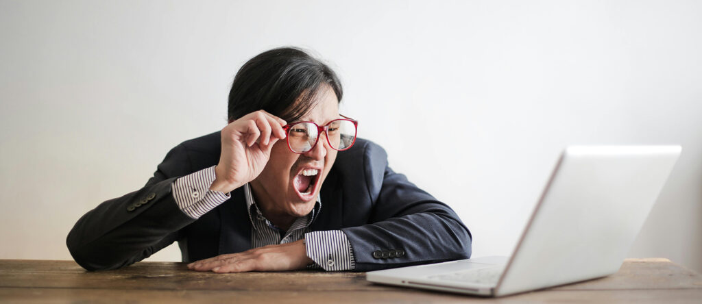A person wearing glasses leans over and screams at an open laptop