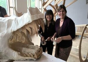 Two women standing behind paper mache of alligator with mouth open