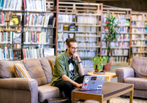 Male college student sitting on couch with laptop in library