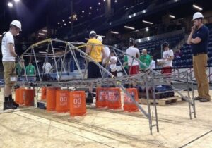 College students wearing hardhats in gym and assessing large model of bridge