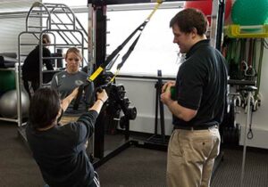 Person using suspended TRX band to do a row with supervision from two others standing nearby