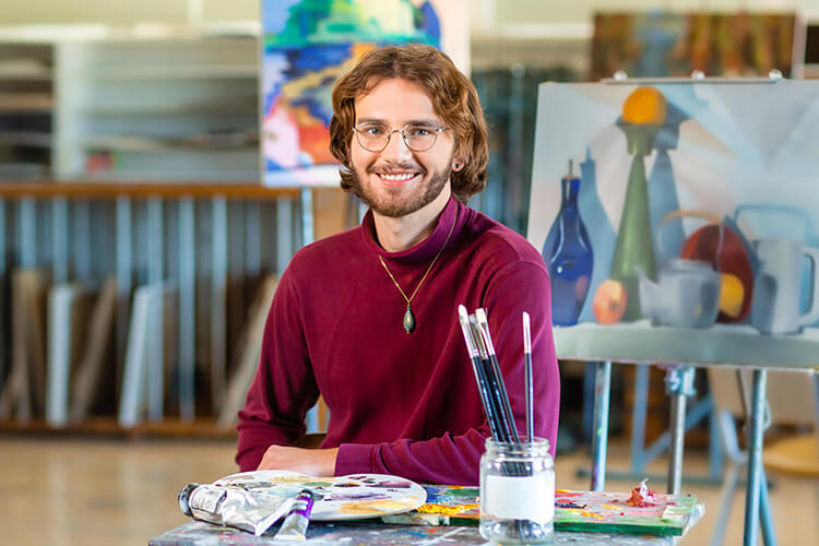 Young man sitting in front of a table with paints and paint brushes with artwork displayed in the background