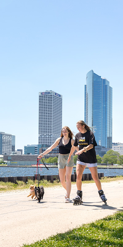 Two girls roller blading and walking a dog with Milwaukee skyscrapers in background