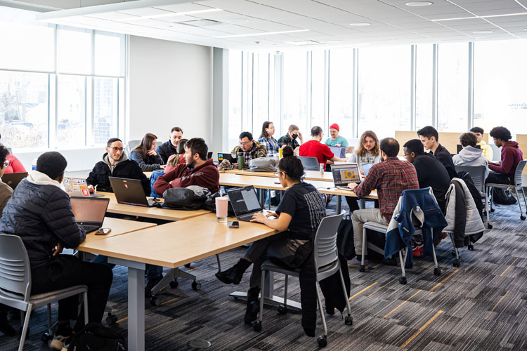 Two college students with backpacks walking on tree-lined campus pathway between buildings.